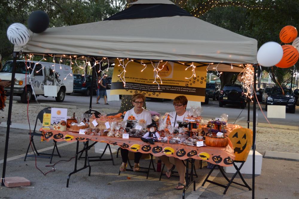 Two women sit behind table with jack-o-lantern-themed table cloth with various food items for sale